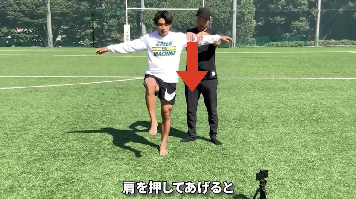 軸足で片足立ちをして腕を広げた選手の肩を、コーチが下に押している